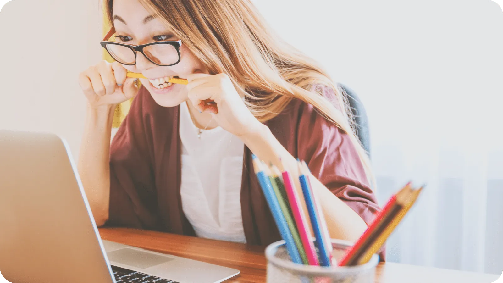 A woman biting a pencil while working at a laptop with a pencil holder nearby